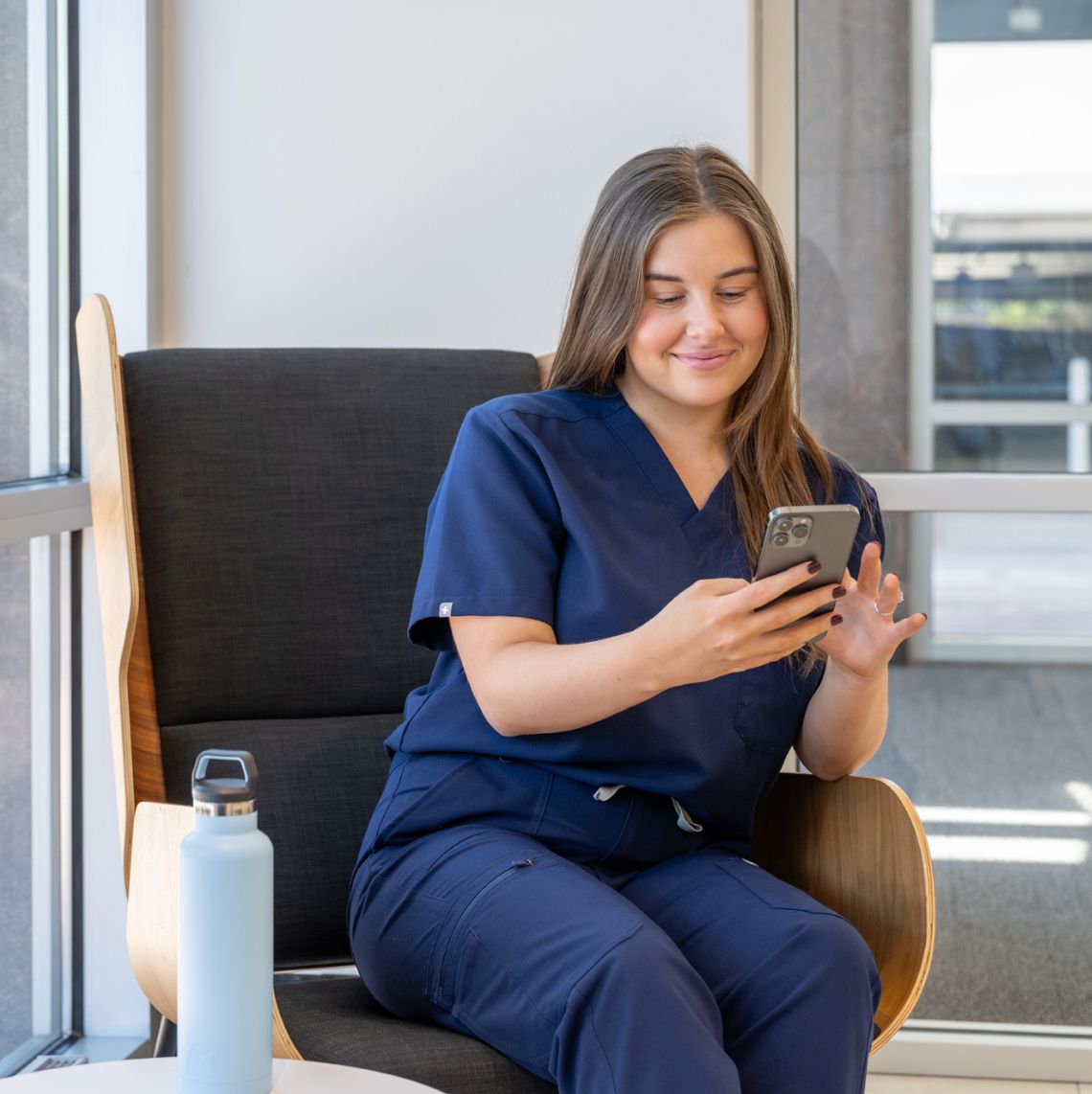 young female healthcare worker in scrubs sitting on chair and using her phone while smiling