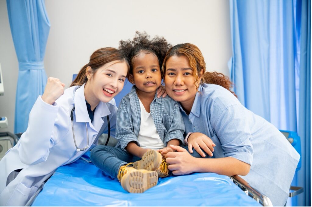 Two nurses standing on opposite sides of a child in hospital bed, cheek to cheek and smiling